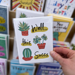 Hand holding a card with cactus and succulent illustrations and text that reads "well this succs" 