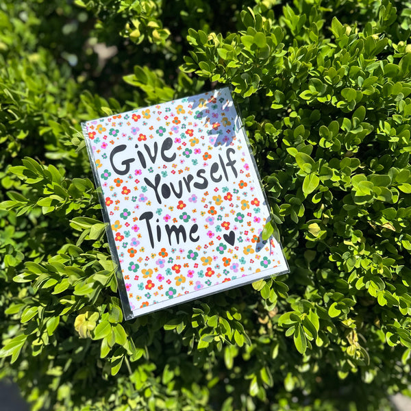 Small card with 'Give Yourself Time' text on a floral pattern, placed on green foliage.